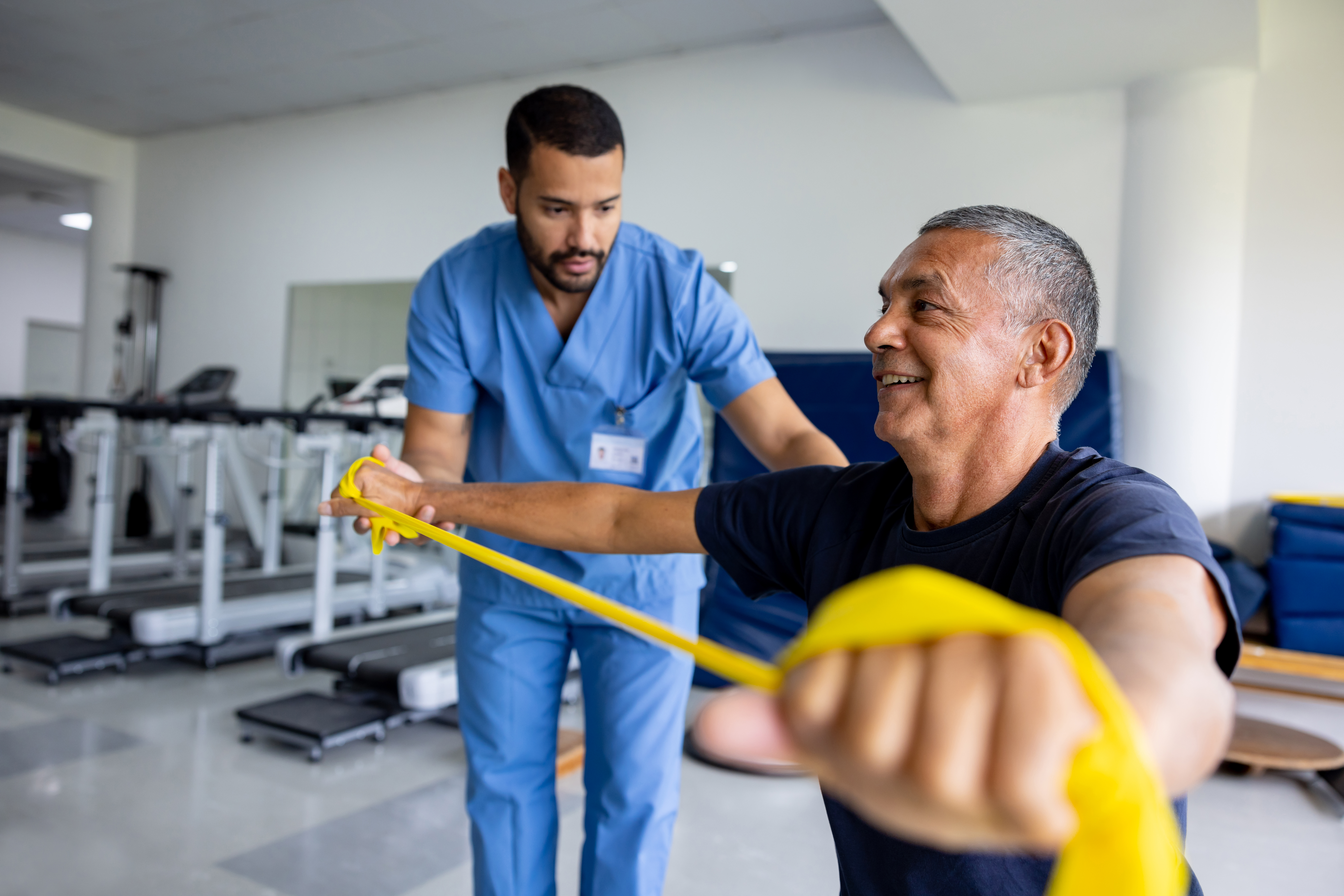 Therapist in blue scrubs guiding elderly patient through resistance band rehabilitation exercise in modern rehab gym
